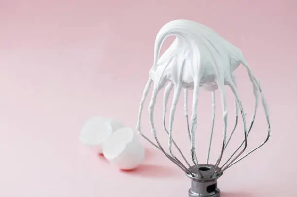 Whipped egg whites - beaten italian meringue on a wire whisk and egg shells on pink background.
