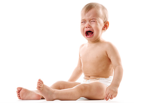 Adorable upset little boy in diaper is sitting and crying on white background.