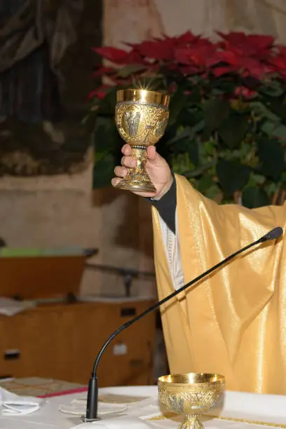 Holy cup containing the blood of Christ in the hand of priest, the sacred grail during ceremony or mass on church or cathedral interior background, close up
