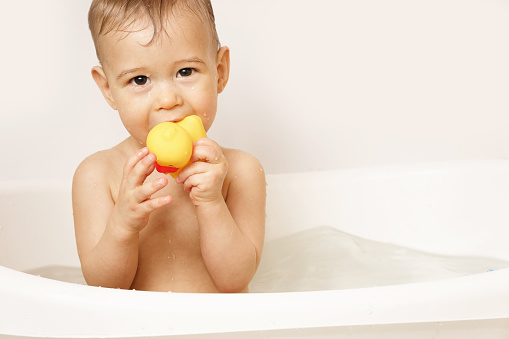 Adorable little boy is putting a rubber duck in his mouth while taking a bath in warm water.