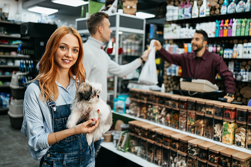 Young adult redhead woman enjoying shopping in pet shop store together with her adorable Shih-Tzu dog. She is smiling and looking at camera.