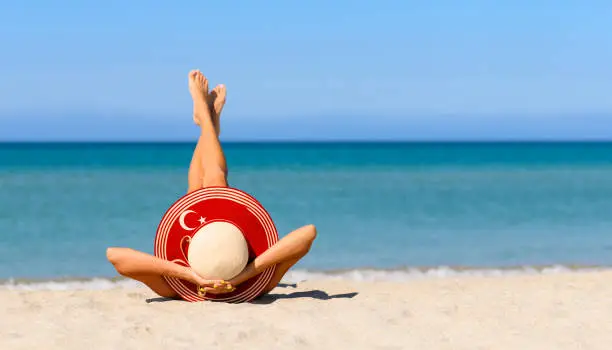 Photo of A slender tanned girl on the beach in a straw hat in the colors of the flag of Turkey. The concept of a perfect vacation in a resort in the Turkey.