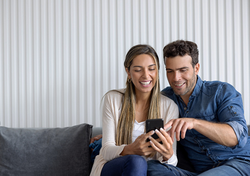 Happy Latin American couple at home laughing while looking at social media on their cell phone - lifestyle concepts
