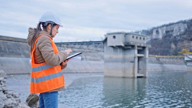 Female engineer working in hydroelectric dam. Ecology orientated. Renewable energy systems. Working day on a water dam with a hydroelectric power plant. Checking the condition of the power equipment, and analysing the data and the results of measurements with a mobile app. reservoir stock pictures, royalty-free photos & images
