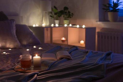 cup of tea with burning candle on wooden tray on bed in bedroom in evening