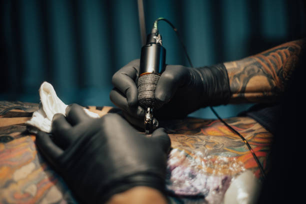 Hands of a tattoo artist wearing black gloves and holding a machine while creating a picture on a man back. Close up of the tattoo machine.