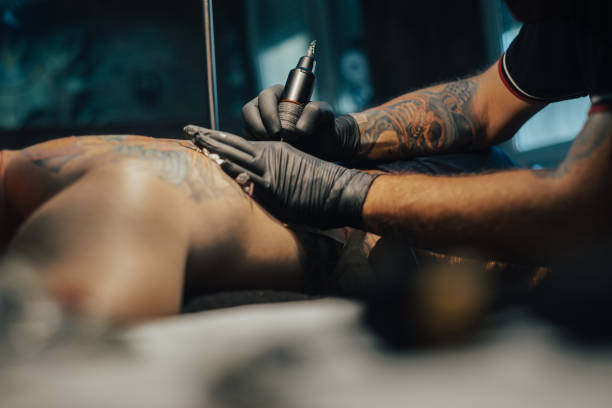 Tattoo artist holding a machine while working in a studio Professional tattoo artist making a tattoo on a man skin while working in a dark tattoo studio. Tattooing. Concept of art and design. black tattoo stock pictures, royalty-free photos & images