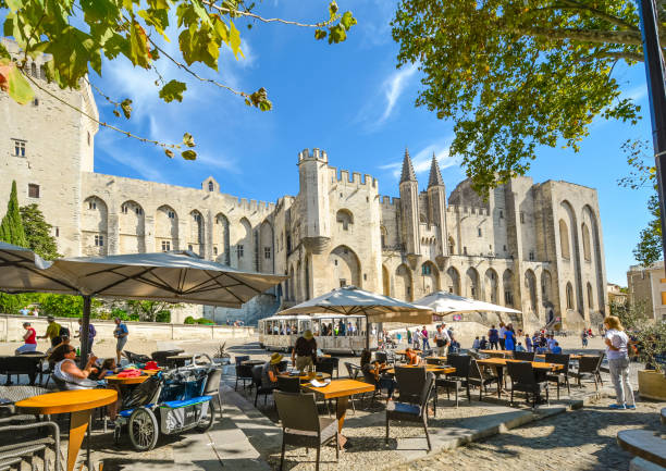 Tourists enjoy a meal at a sidewalk cafe in front of the medieval Pope's Palace in the Provence region of Avignon France as a tourist tram drives by on a sunny day Tourists enjoy a meal at a sidewalk cafe in front of the medieval Pope's Palace in the Provence region of Avignon France as a tourist tram drives by on a sunny day avignon france stock pictures, royalty-free photos & images