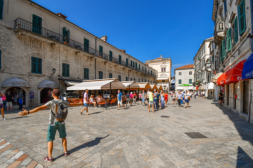 A man poses for a photo with outstretched arms in the Piazza or Square of the Arms in the ancient city of Kotor, Montenegro as tourists walk by the cafes and shops