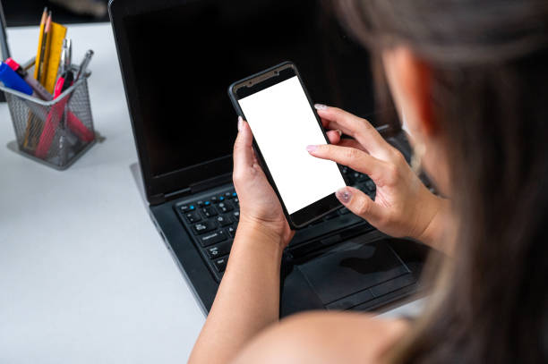 istock Top view mockup image of young woman hands holding blank mobile phone while using laptop with blank black desktop screen on table 1370611969