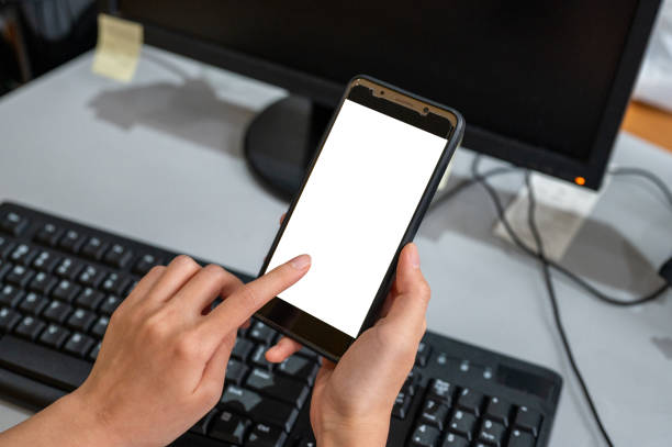 istock Top view mockup image of woman hands holding blank mobile phone while using computer with blank black desktop screen on table 1370611980