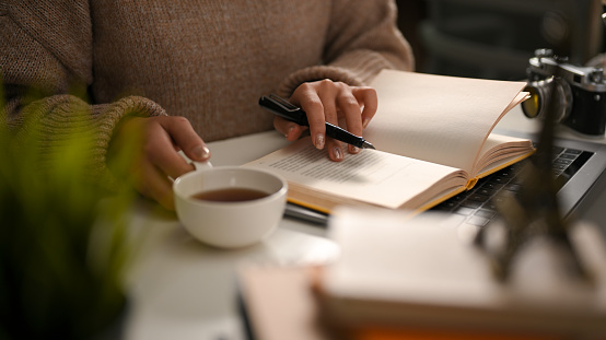 A female reading a book or writing a diary and sipping hot tea in cozy home workspace. leisure and hobby concept