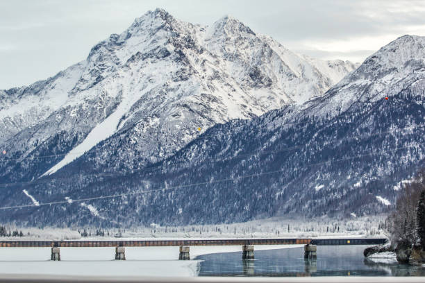Traveling through Alaska has various means. One way tourists and locals alike choose to travel is through the rail system. Visitors will cross over many miles and see stunning sights. One place they can view on the way North from Anchorage takes the traveler across the Knik River. The railroad bridge crosses right over the river and provides a stunning view of the mountains. On this frigid day the bridge made for a spectacular view.