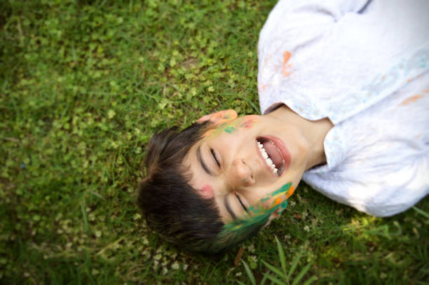 白い色のクルタの幸せな明るい若い男の子は、目を閉じて歯を微笑んでいる、緑の草の上に横たわっているホーリーパウダーカラーで描かれた顔 - green tika ストックフォトと画像