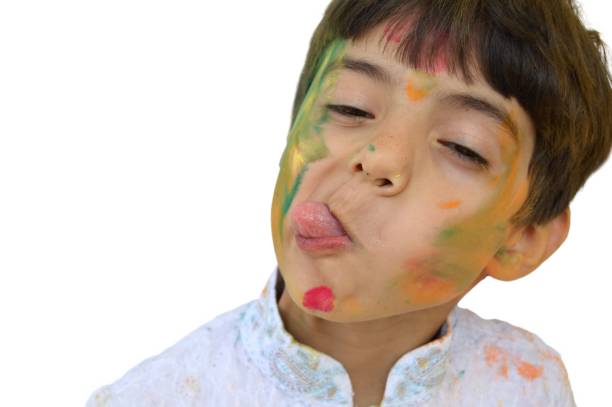 cut out of a horizontal photograph of a young boy in white coloured kurta teasing by his tongue sticking out, face and hair painted with holi powder colours, making an ugly tilted funny  face - making a face mischief sticking out tongue isolated on red imagens e fotografias de stock