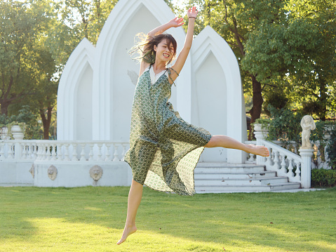 Portrait of young Asian woman jump into air with open legs and arms in sunny day, beautiful Chinese girl with green hairs in green dress dancing in garden carefreely, full length shot.