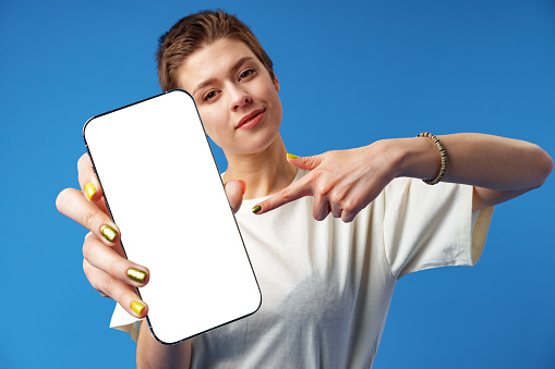 Portrait of a young woman showing blank screen mobile phone while standing over blue background, close up