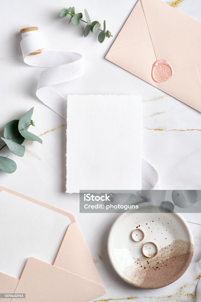 Elegant wedding stationery top view. Flat lay blank paper card mockup, pink envelopes, eucalyptus branches, golden rings on marble background. Office Supply Stock Photo