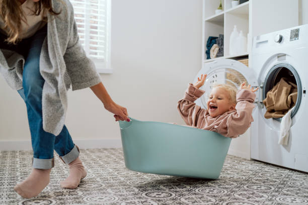 mom is playing with daughter who is sitting in laundry bowl little girl wants to spend time with woman and help with household chores mother drags her daughter around laundry room for fun in bowl - de was doen stockfoto's en -beelden