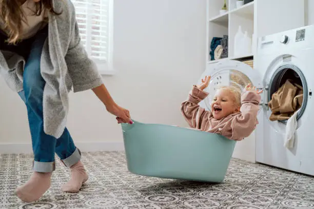 Photo of Mom is playing with daughter who is sitting in laundry bowl little girl wants to spend time with woman and help with household chores mother drags her daughter around laundry room for fun in bowl