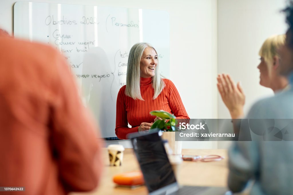 Barcelona businesswoman presenting objectives for new year Personal perspective of mid 50s executive smiling and looking away as she receives applause from colleagues for her ideas at informal meeting. Office Stock Photo