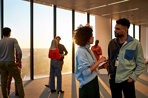 Casually dressed men and women of mixed age range standing in unfurnished modern environment with full length windows and view of city at sunset.