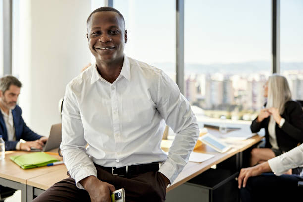 Portrait of relaxed Black businessman in meeting room Front view of mid 20s professional in button down shirt and pants sitting on end of table, holding phone, and smiling at camera with associates in background. project manager stock pictures, royalty-free photos & images