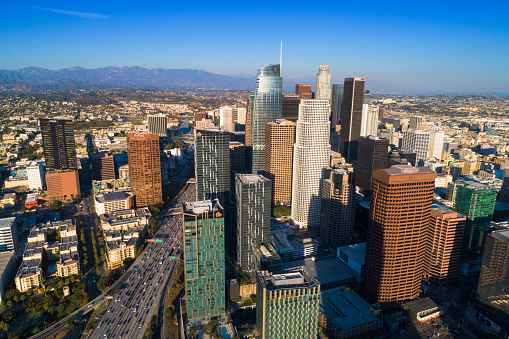 Downtown Los Angeles skyline aerial view during the late afternoon, with the 110 freeway with traffic on the bottom left and the San Gabriel Mountains in the far distance.