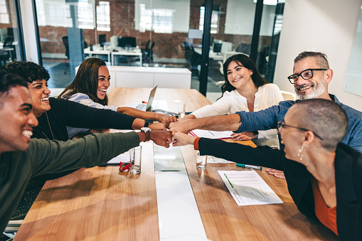 Team of happy businesspeople bringing their fists together while sitting in a boardroom. Diverse group of colleagues smiling cheerfully during a meeting in modern workplace.