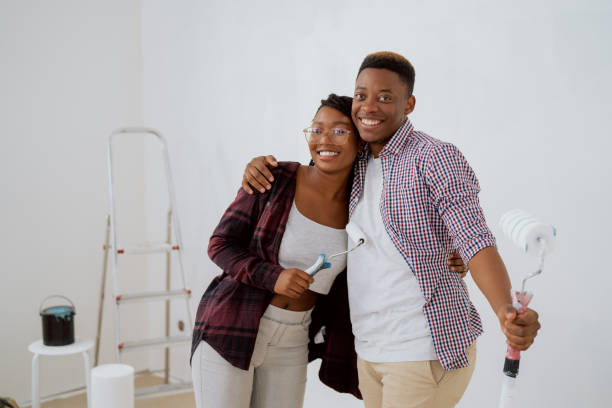 istock Handsome dark-skinned boy embraces beautiful smiling woman hold wall paint rollers in hand, couple hugs, together renovate new apartment, refresh white wall color 1370659763