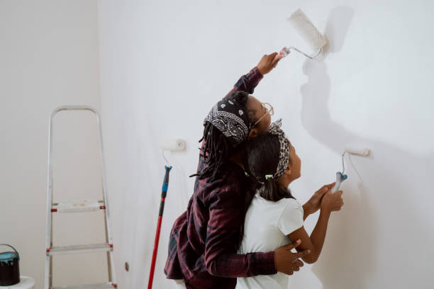 maman occupée passe du temps avec sa fille joyeuse, l’engage pour l’aider, ensemble ils peignent les murs de l’appartement en rénovation, appliquent de la peinture blanche avec des rouleaux - home improvement home interior painting family photos et images de collection