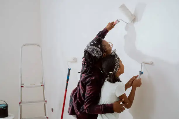 Photo of Busy mom spends time with cheerful daughter, engages her to help, together they painting the walls of the apartment under renovation, apply white paint with rollers