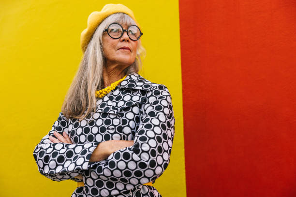 Retired woman looking away thoughtfully against a colourful wall Retired woman looking away thoughtfully while standing against a red background. Pensive elderly woman reflecting on memories of the past. Stylish senior woman wearing colourful clothing. multi coloured glasses stock pictures, royalty-free photos & images