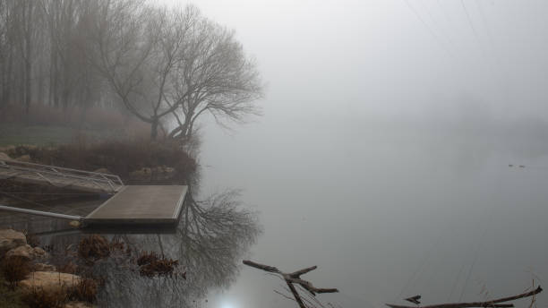 On the bank of a river, a wooden pier overlooks the water in the cold morning fog, the atmosphere is cold and with little visibility.
