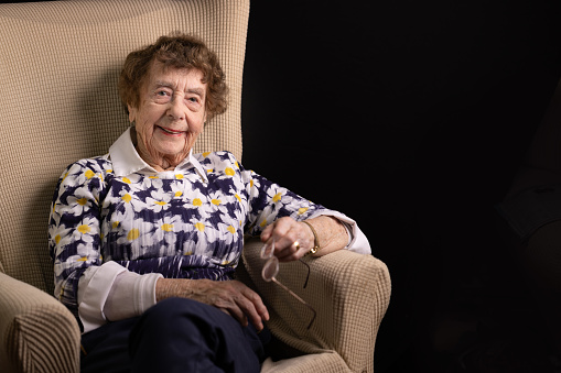 Portrait of an active, cheeky, happy and healthy 94 year old great grandmother sat in her living room chair smiling at the camera. Shot against a black background.