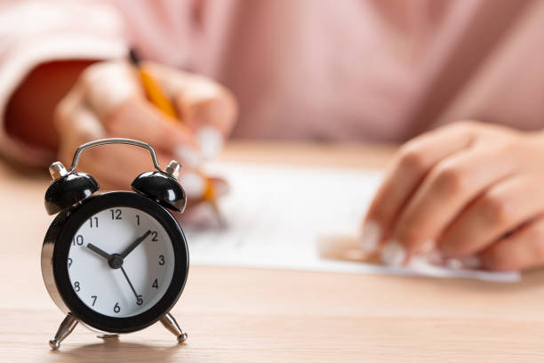 istock alarm clock with time running out. Students hands to take exams, write examination room with pencil holding on optical form of standardized test with answers 1370669743