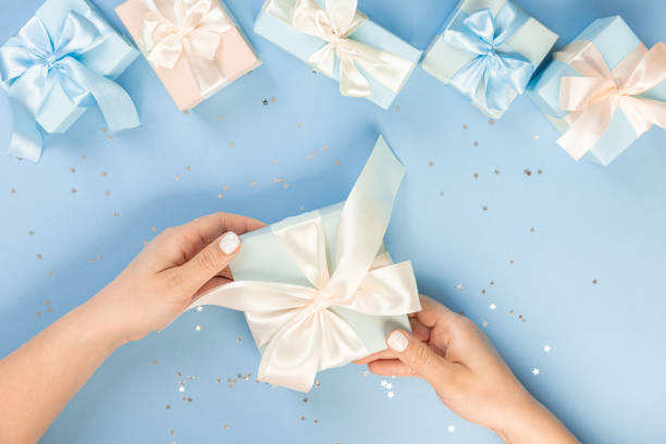 istock Flat lay of women's hands holding a gift wrapped and decorated with a bow on a blue background with silver sequins with copy space part of the table is surrounded by several festively wrapped gifts 1370670708