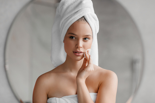 Beauty routine. Pretty young woman cleaning her face with cotton pad at home, looking at mirror, standing with towel on head after bath, reflecting in mirror