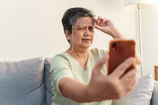 Mujer asiática mayor con presbicia, quitándose los anteojos mientras lee el mensaje desde el teléfono inteligente. photo