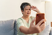 Senior Asian woman with presbyopia, taking off eyeglasses while reading message from smart phone.