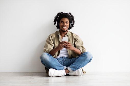 Gadgets For Leisure. Smiling Black Guy Relaxing With Smartphone And Wireless Headphones On Floor, Cheerful Young African American Man Listening Music Online And Smiling At Camera, Copy Space