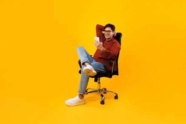 Photo of Man Using Smartphone Sitting In Chair Texting Over Yellow Background