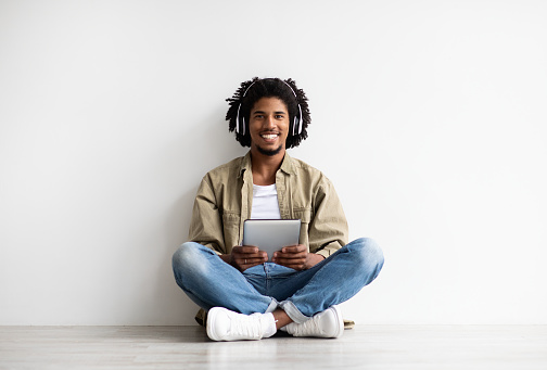 Modern Technologies. Happy Black Guy In Wireless Headphones Using Digital Tablet While Relaxing On Floor Near White Wall, Cheerful Young African American Man Resting With Gadgets At Home, Copy Space