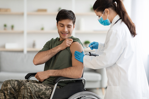 Positive young man in camouflage uniform soldier in wheelchair getting immunization against coronavirus at clinic, nurse in protective workwear making shot in shoulder for military man, copy space