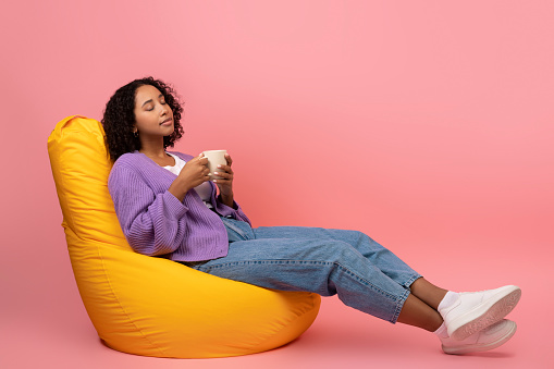 Millennial black female drinking coffee, relaxing in bean bag chair on pink studio background, copy space. Calm young African American woman enjoying weekend with hot drink