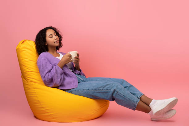 Millennial black female drinking coffee, relaxing in bean bag chair on pink studio background, copy space Millennial black female drinking coffee, relaxing in bean bag chair on pink studio background, copy space. Calm young African American woman enjoying weekend with hot drink bean bag stock pictures, royalty-free photos & images