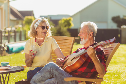 Cheerful senior couple having an outdoor lunch in the backyard, sitting at the table, drinking beer and lemonade, playing the guitar, singing and relaxing on a sunny summer day