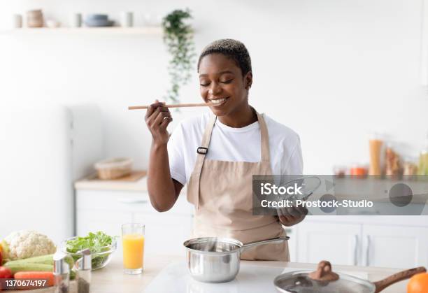 Smiling Millennial African American Woman In Apron Cooking At Home And Tasting Dish In Kitchen Interior Stock Photo - Download Image Now