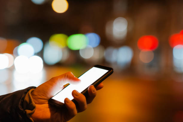 istock Smartphone with white screen in human hand, copy space in darkness with bright bokeh lights. Using phone in the city at night 1370677194