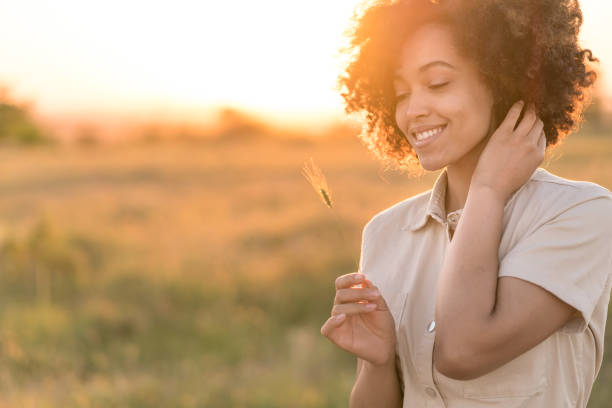 Portrait of beautiful african american woman holding a grass straw, smiling and looking at the camera in front of grass field during sunset. Outdoor portrait of a smiling multiracial girl. Happy cheerful girl laughing in the nature retrato de una hermosa mujer afroamericana sosteniendo una pajita de hierba, sonriendo y mirando a la cámara frente al campo de hierba durante la puesta de sol. retrato al aire libre de una sonriente chica multirracial. feliz chica alegre riendo en la nat